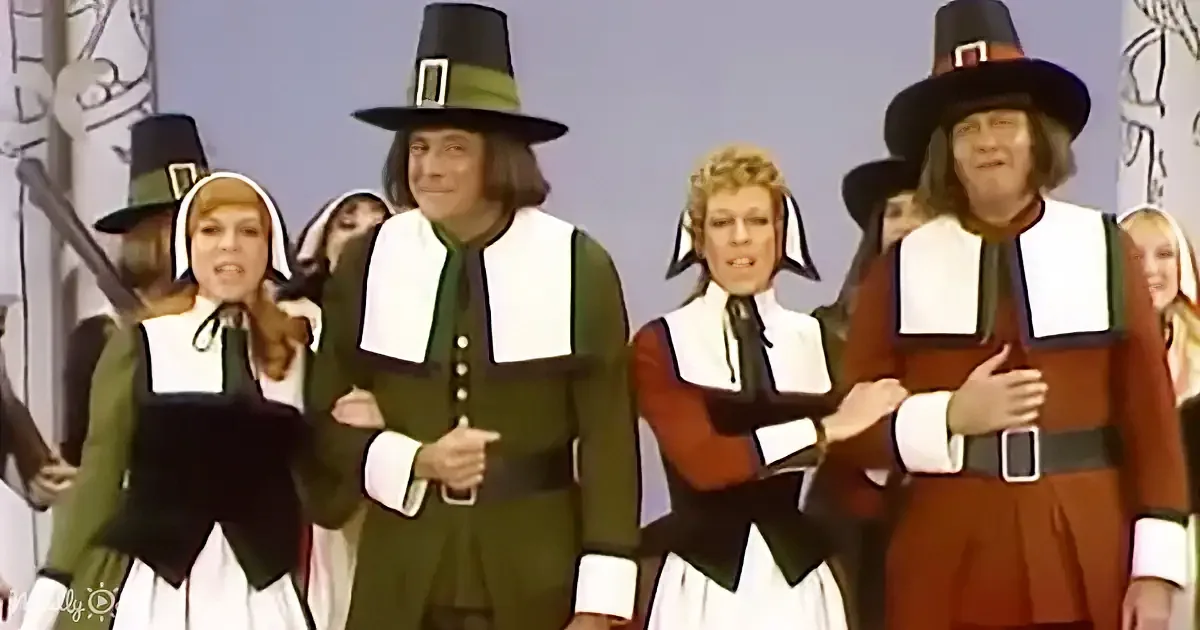 A group of people dressed in Pilgrim costumes, including hats and aprons, are singing or performing.