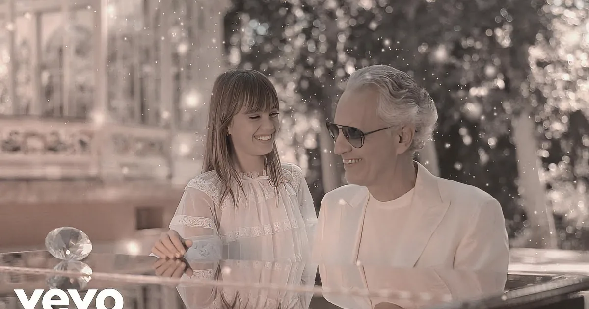 A young girl in white lace dress stands behind an older man in white, both in a snowy setting.