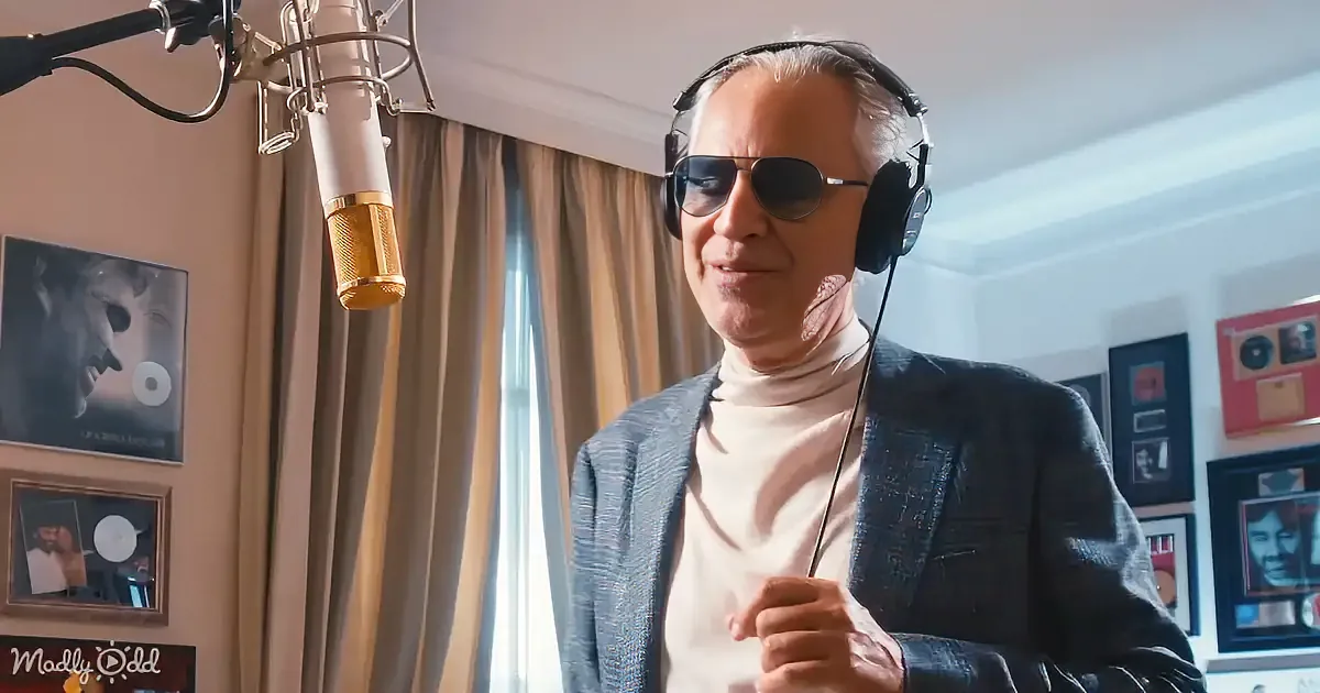 A man wearing headphones and sunglasses stands in front of a microphone in a recording studio.