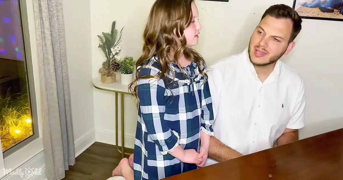 A young girl in a blue plaid dress stands next to a man in a white shirt, both looking at each other in a cozy room.