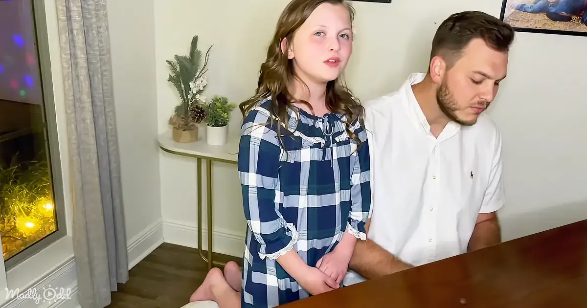 A young girl in a blue plaid dress stands beside a man in a white shirt, both looking down at a table.