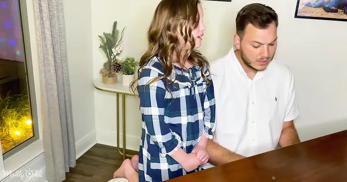 A young girl in a plaid dress stands beside a man in a white shirt at a table, with a cozy room background.