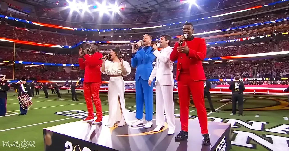 A group of five performers in colorful suits sing on a stage at a large stadium.
