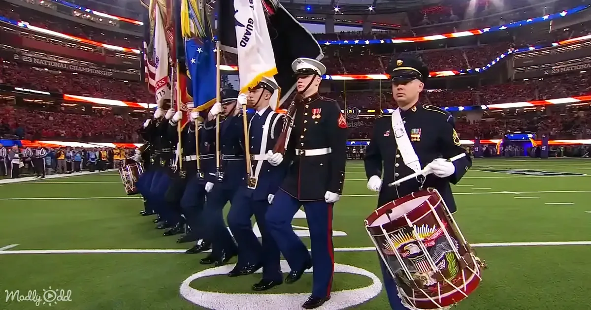 A military band in uniform marches on a football field with flags and drums.