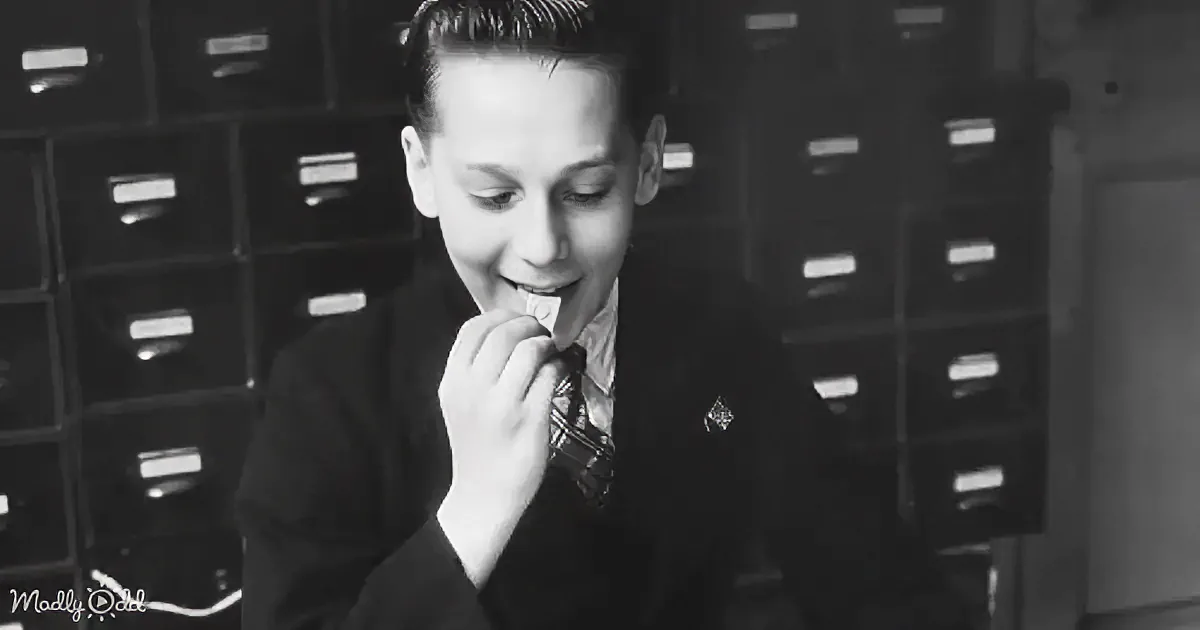 A boy in a suit and tie is eating something, with a filing cabinet in the background.