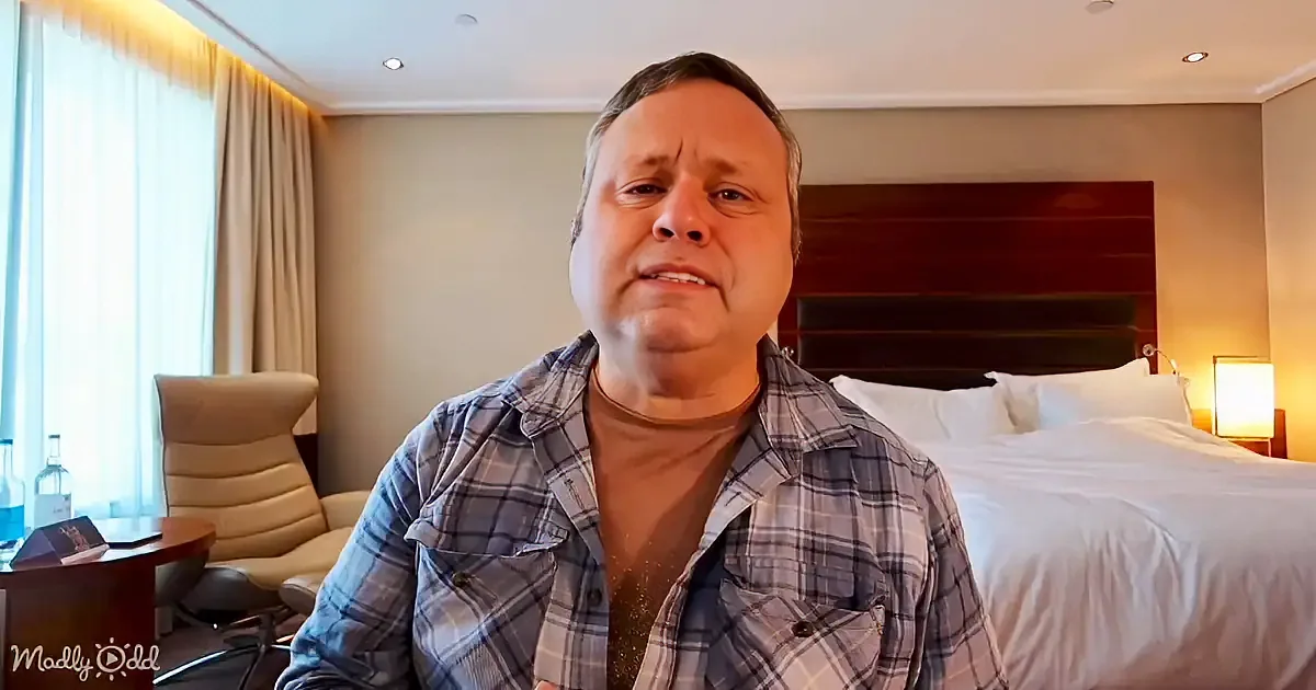 A man in a plaid shirt sits in a hotel room with a bed, chair, and lamp in the background.
