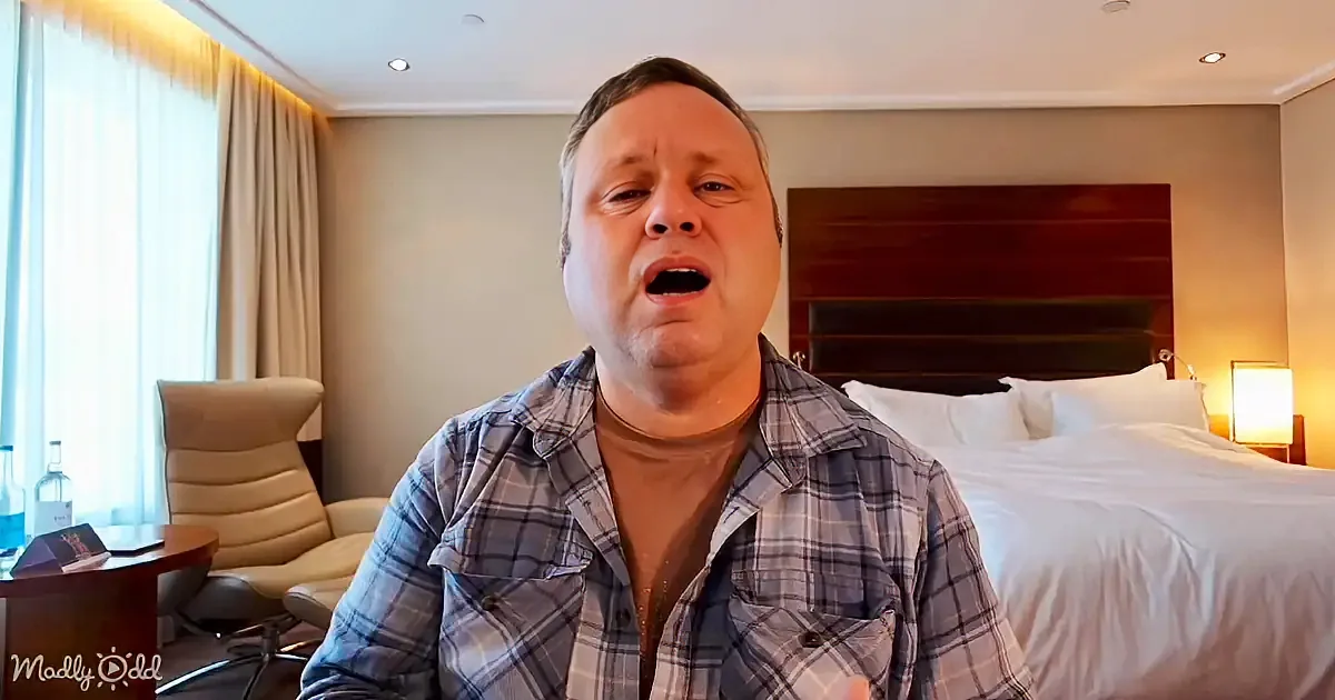 A man in a plaid shirt sits in a hotel room, mouth open, with a bed and chair in the background.