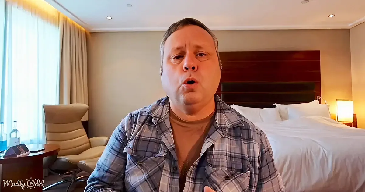 A man in a plaid shirt sits in a hotel room, speaking, with a bed and chair in the background.