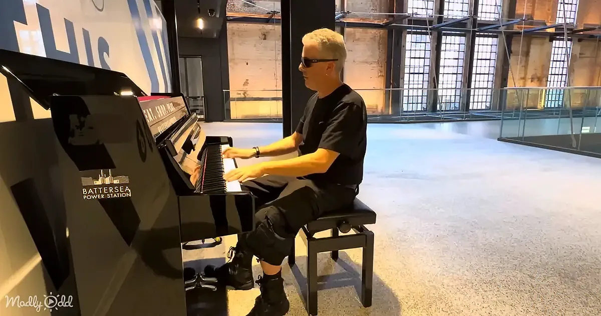 A man in black plays a piano at Battersea Power Station, wearing sunglasses and black attire.