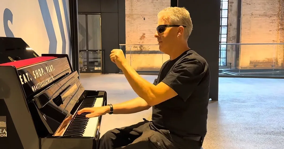 A man in sunglasses plays a piano in a modern, spacious room with large windows.