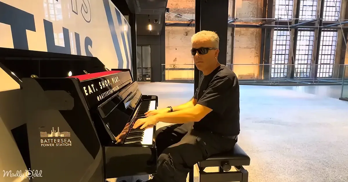 A man in sunglasses plays a piano in a spacious, industrial-style room with large windows.