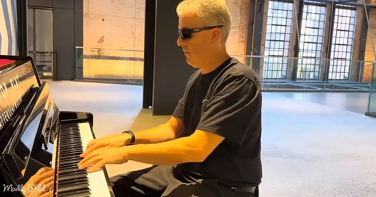 A person wearing sunglasses and a black shirt plays a piano in a spacious room with large windows.