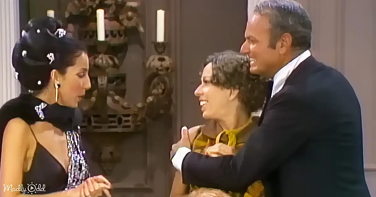A man in a suit embraces a woman in a yellow dress, while another woman in a black dress looks on.