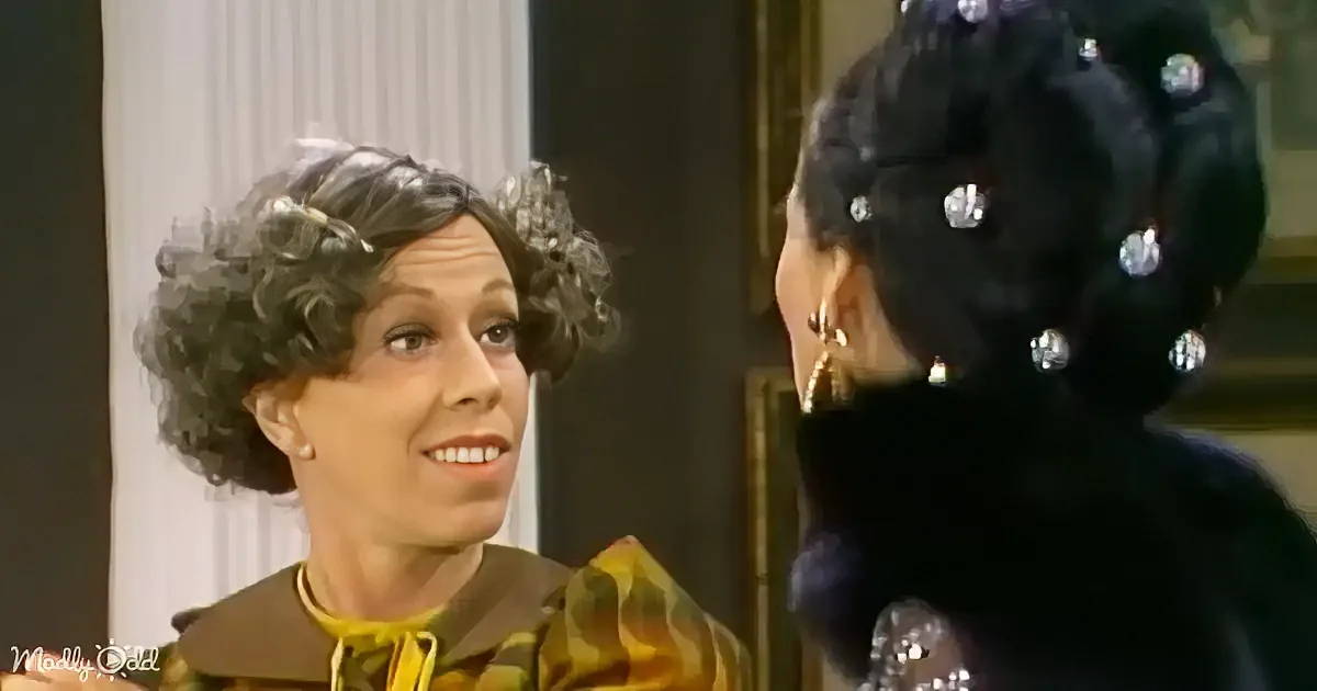 A woman with curly hair and a yellow and brown outfit smiles at another woman in a black dress with sparkling details.