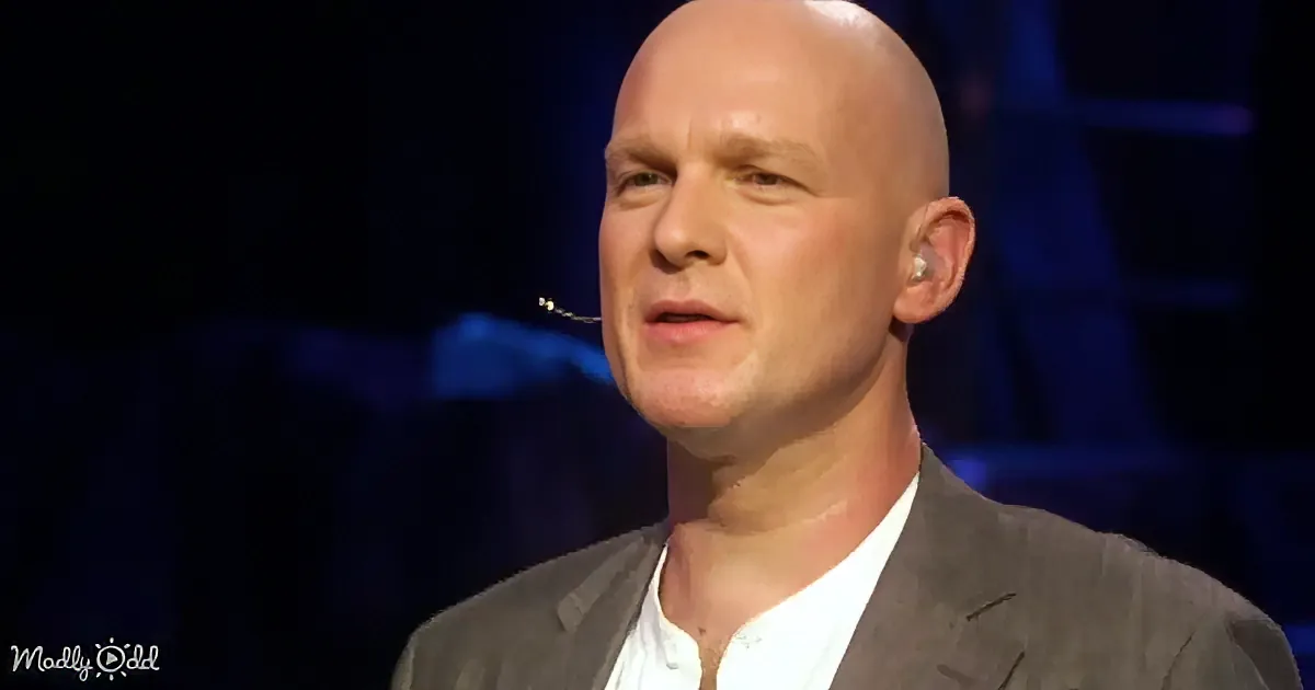 A bald man in a suit with a microphone headset, speaking on stage.