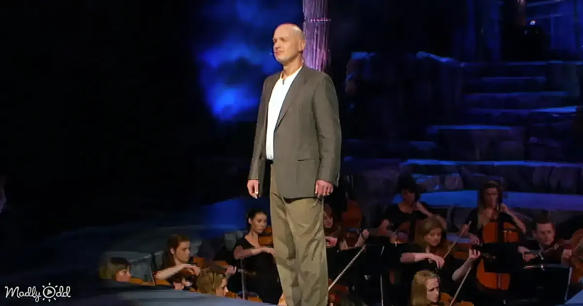 A man in a suit stands on stage with an orchestra behind him, illuminated by blue stage lights.
