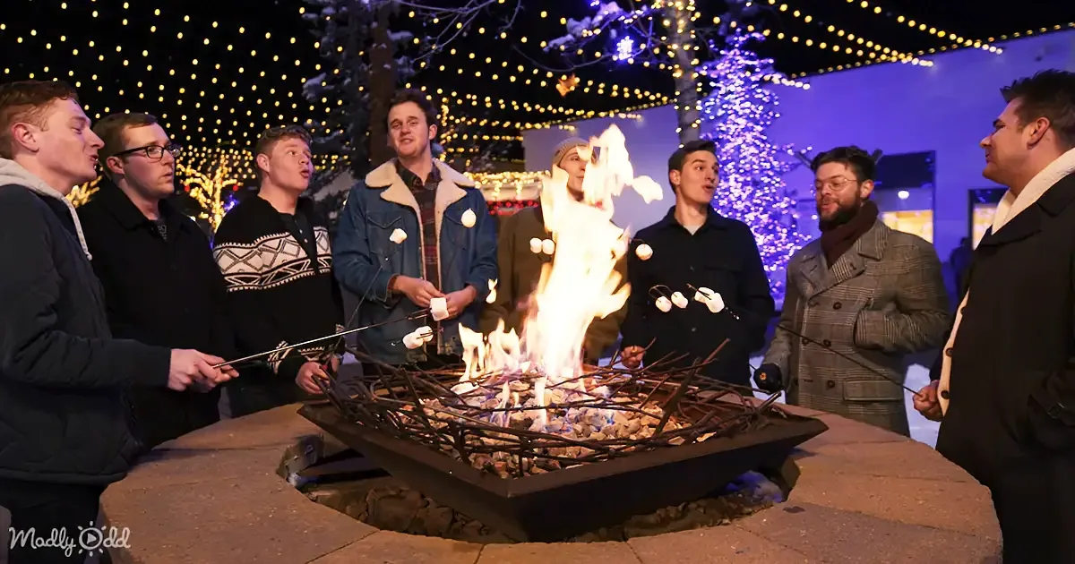 A group of people roast marshmallows around a fire pit under festive lights.