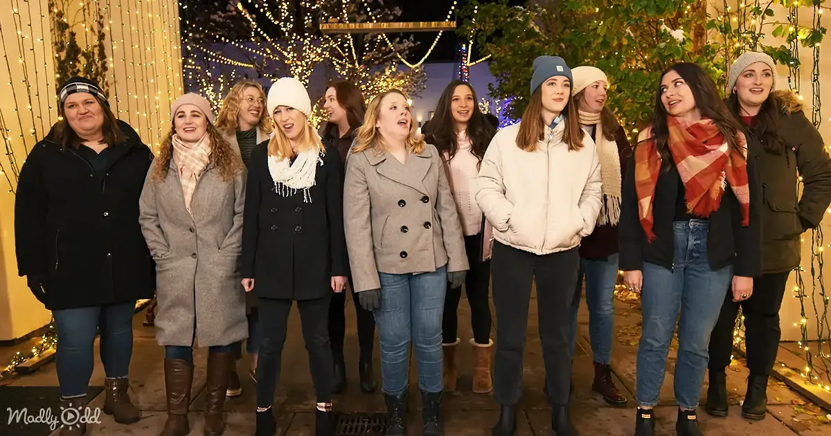 A group of women in winter attire stands together, smiling and looking up, surrounded by festive lights.