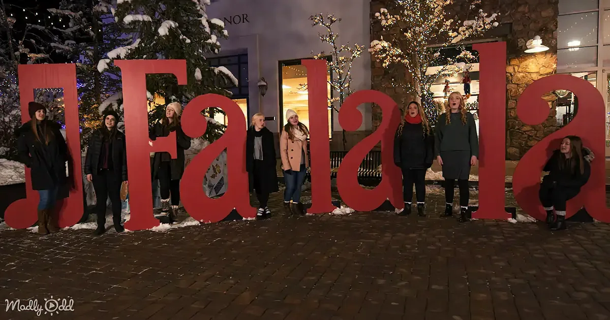 Women sing behind giant "Fa la la" letters in snowy, festive setting.