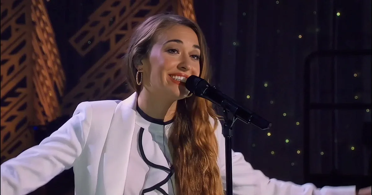 A woman with long hair sings into a microphone, wearing a white outfit, with a dark background and a spotlight effect.
