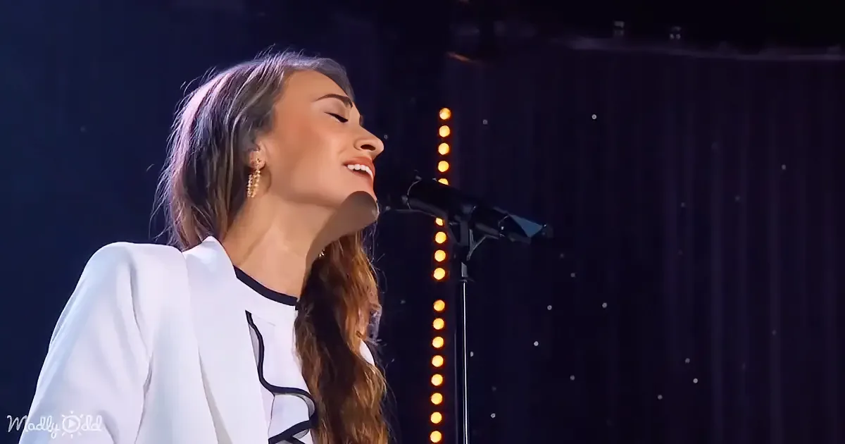 A woman in a white blazer sings passionately into a microphone on stage with a string of lights in the background.