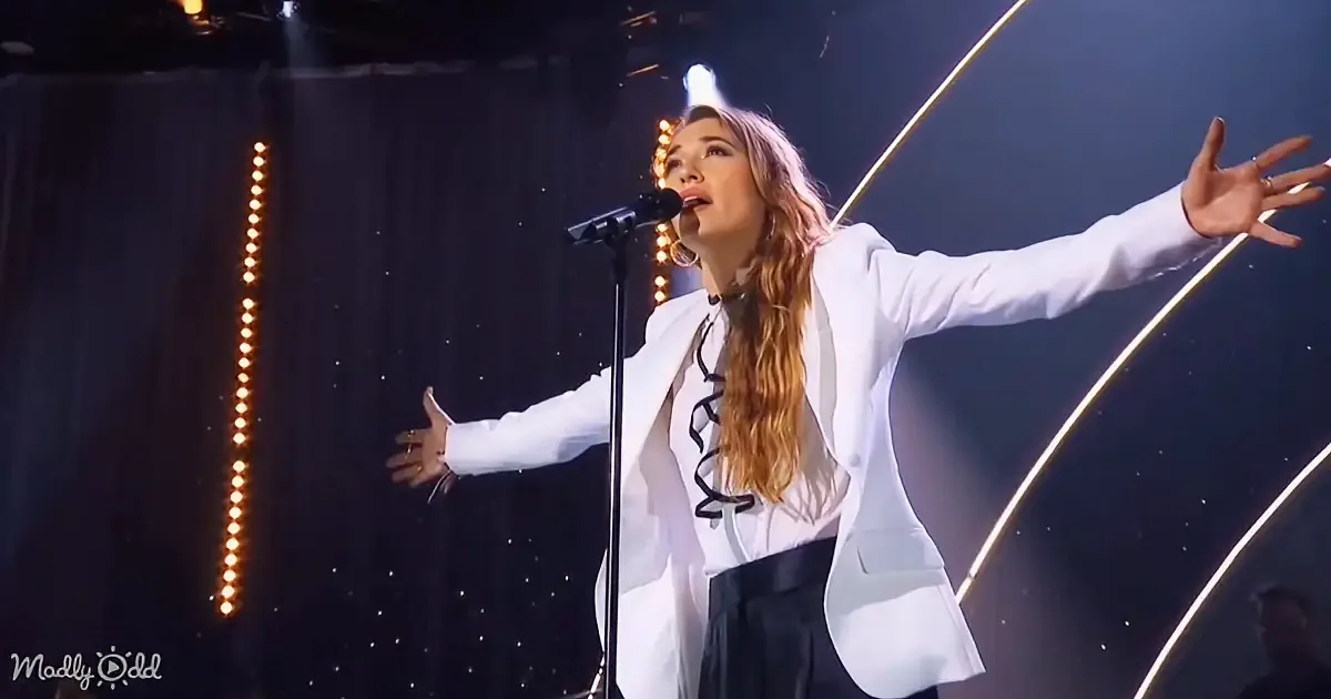 A woman in a white blazer sings passionately on stage with arms outstretched, surrounded by stage lights.