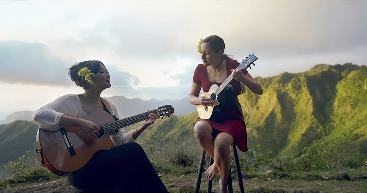 Two women play guitar and ukulele on a mountain top, surrounded by greenery.