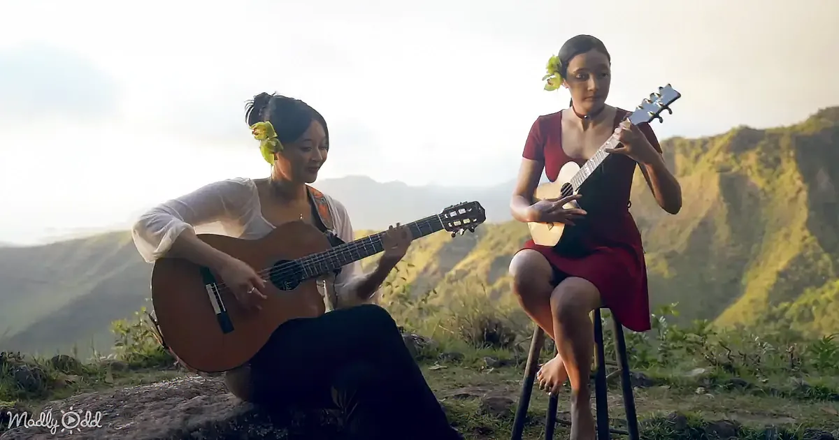 Two women play guitar and ukulele on a mountain top, surrounded by lush greenery.