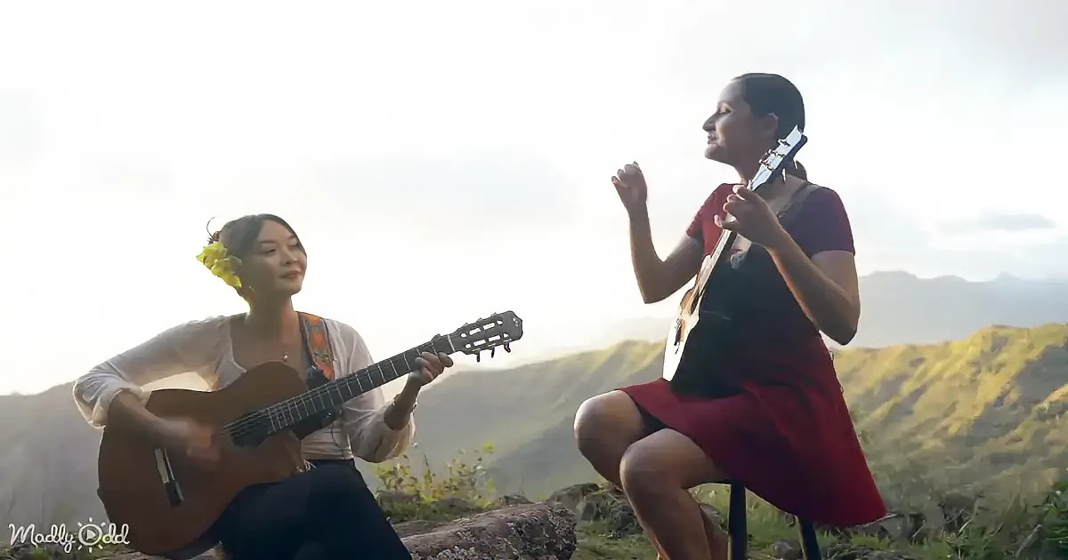 Two women play guitars outdoors, one in white, the other in red, against a scenic mountain backdrop.