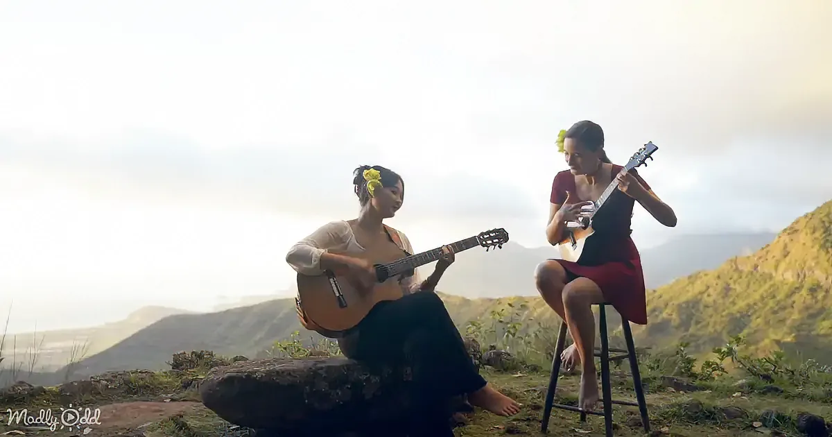 Two women play guitars on a hilltop, surrounded by nature, under a cloudy sky.
