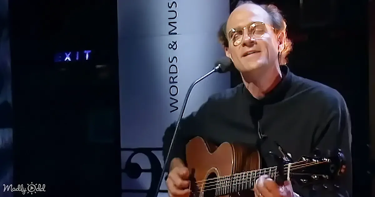 A man in glasses plays an acoustic guitar on stage, with a microphone and "EXIT" sign visible in the background.