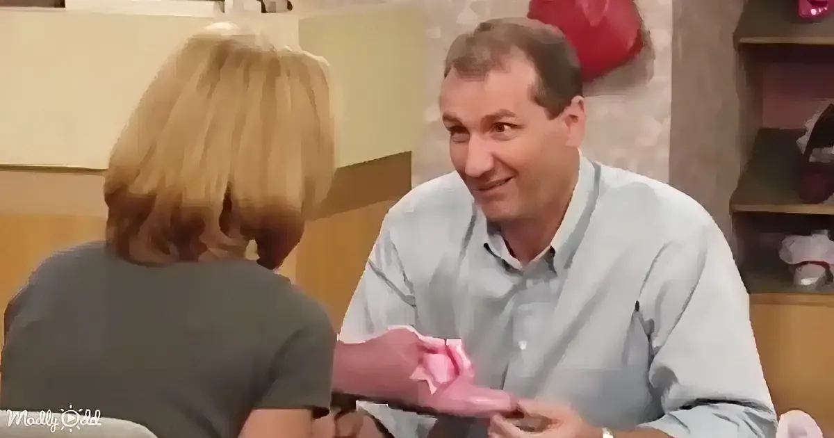 A woman in a green top sits on a couch, holding a pink object, while a man in a white shirt listens.