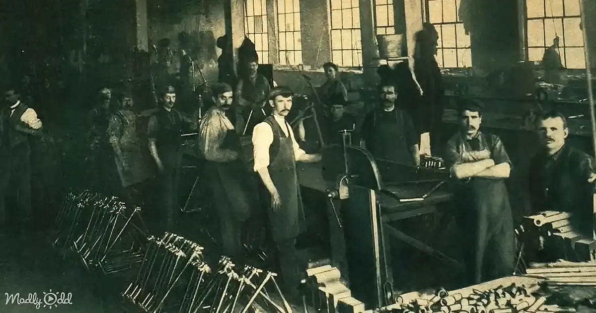 Men in a workshop with tools and machinery, some wearing aprons, in a dimly lit industrial setting.
