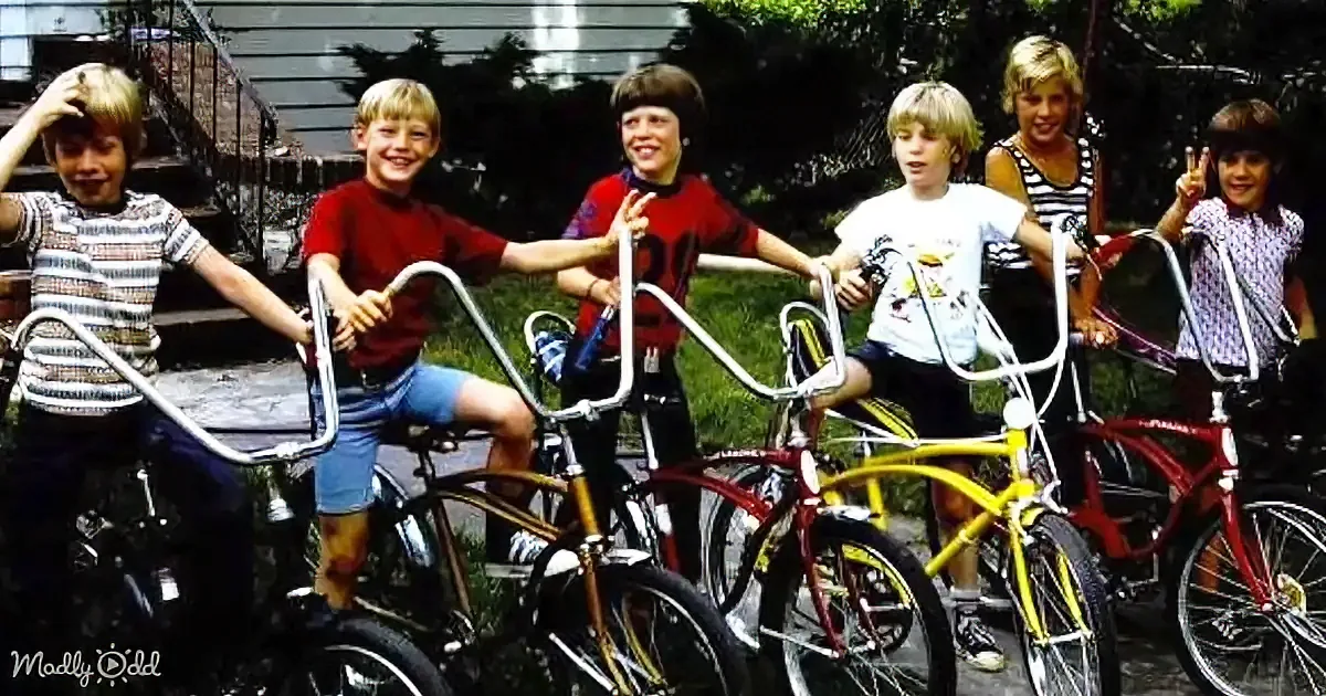 Six children on bicycles smile and pose outdoors in a suburban setting.