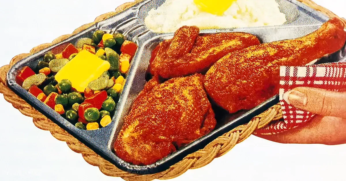 A hand holds a tray with fried chicken, mashed potatoes, vegetables, and butter.