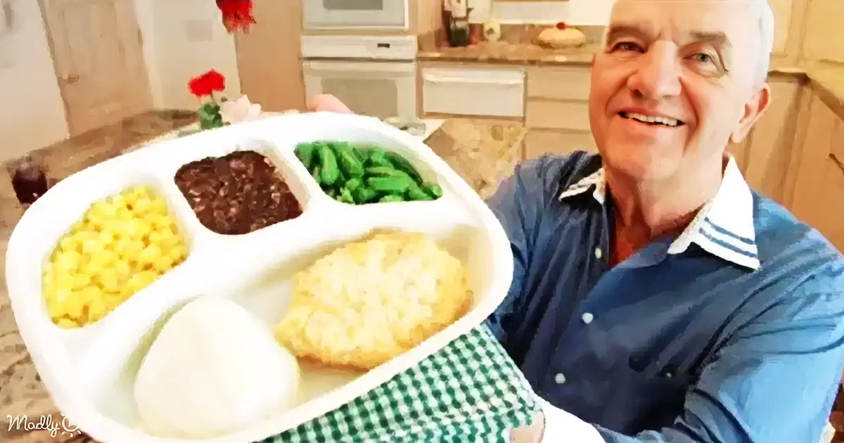 A man holds a tray with corn, beans, green beans, a biscuit, and a white food item, smiling in a kitchen.