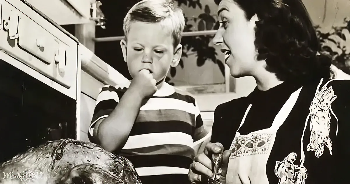 A young boy in a striped shirt holds his finger to his mouth, looking at a woman in an apron who is holding a spoon.
