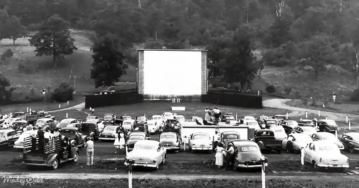 A vintage drive-in movie theater with cars parked in rows, people mingling, and a large screen displaying a bright image.