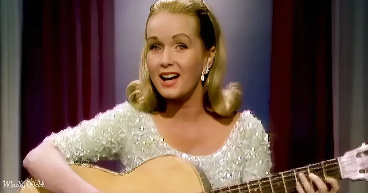 A woman in a sparkly dress plays a guitar and sings on stage with red curtains behind her.