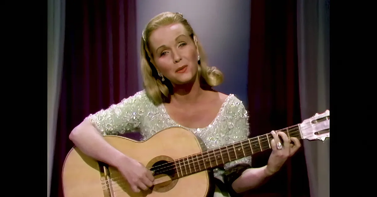 A woman in a sparkly white dress plays a guitar on stage with a purple curtain backdrop.