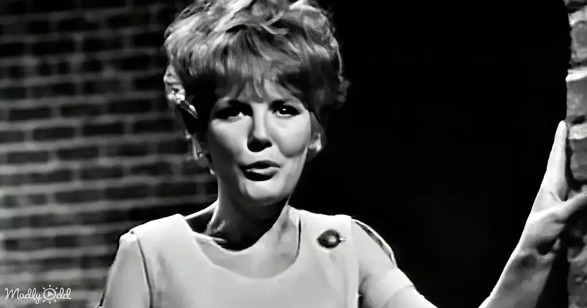 A woman with short, curly hair in a light-colored dress stands against a brick wall.