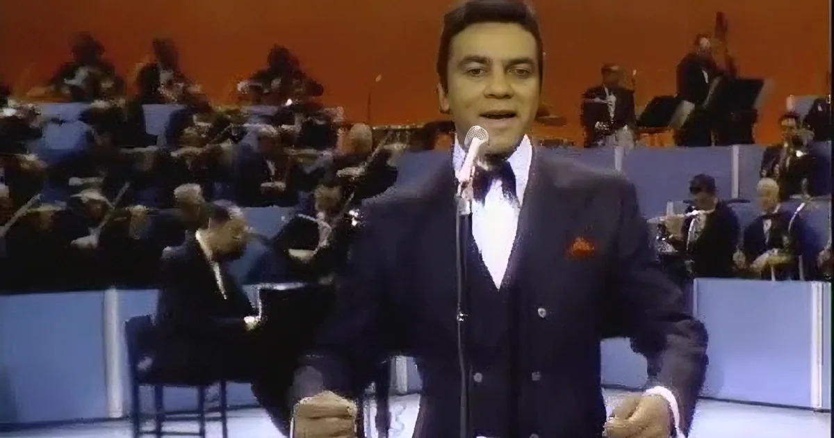 A man in a suit sings into a microphone on stage, with musicians in the background.