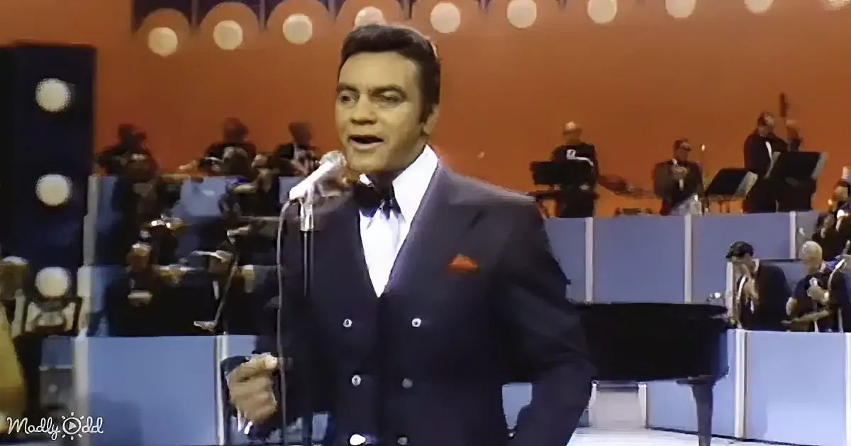 A man in a suit sings into a microphone on stage with an orchestra behind him.