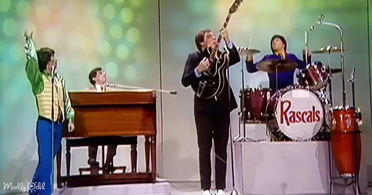 A man in a suit plays guitar while another in a green vest sings into a microphone on stage.