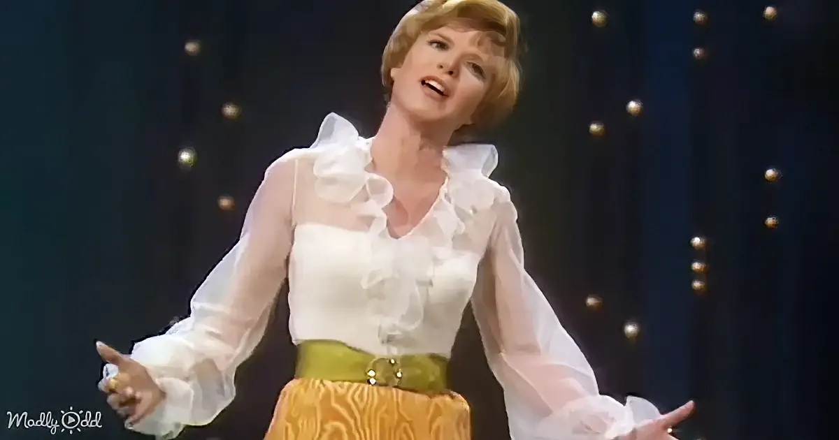 A smiling woman with short blonde hair, wearing a white ruffled top, against a dark background.
