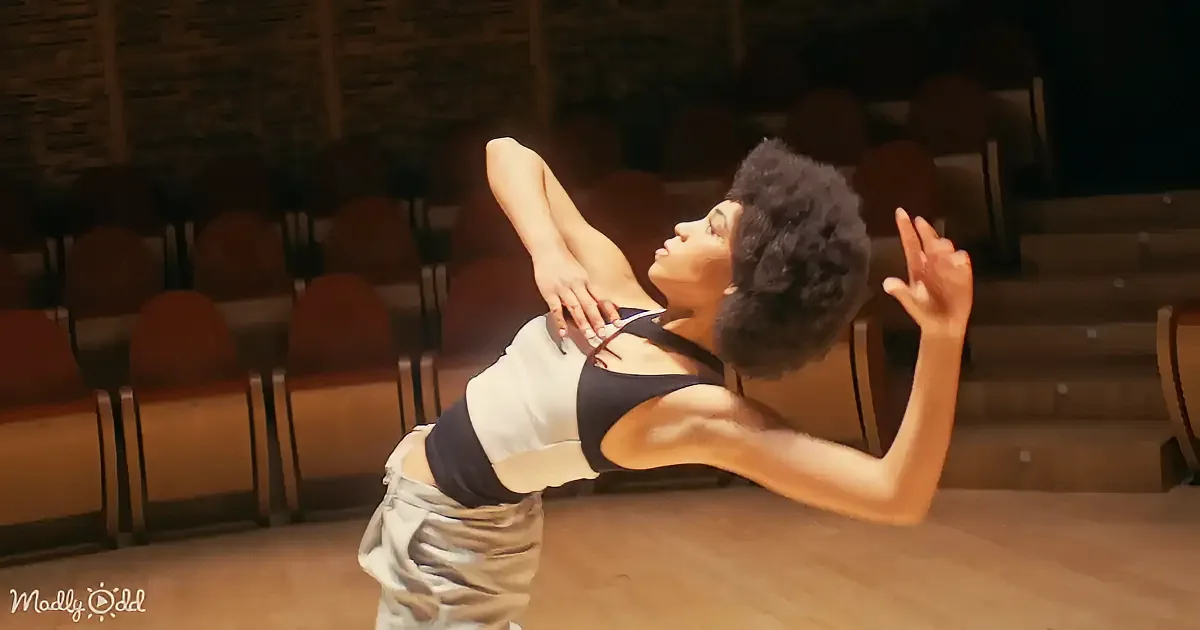 A dancer in a white and black outfit extends their arms gracefully on a stage with wooden chairs in the background.