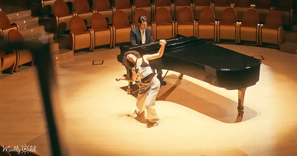 A woman in white pants and a black top dances gracefully in front of a man playing a grand piano in an empty auditorium.