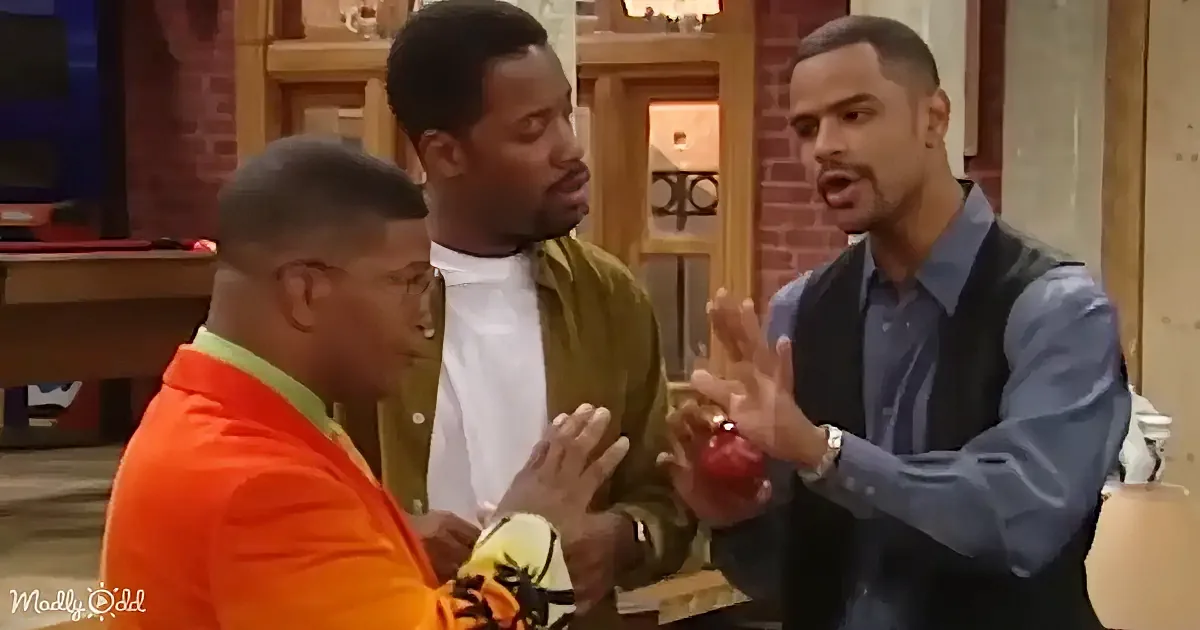 A man in a blue shirt holds a red apple, gesturing while talking to two others in a room.