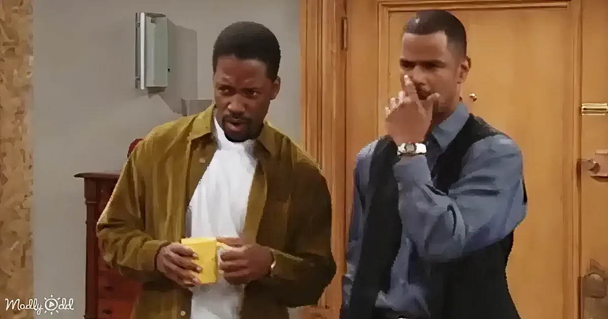 Two men stand in a room; one smokes, the other holds a yellow object, both appear to be in a casual setting.