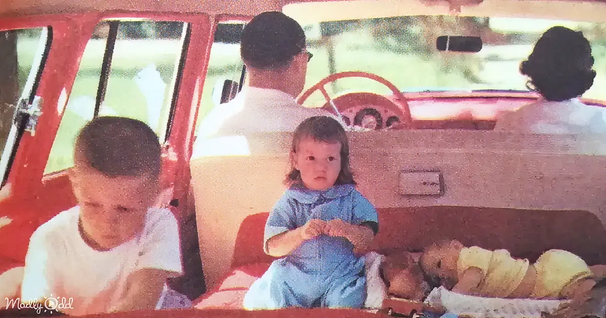 A family sits in a vintage red car, with a baby in the front seat and two children in the back, enjoying a road trip.